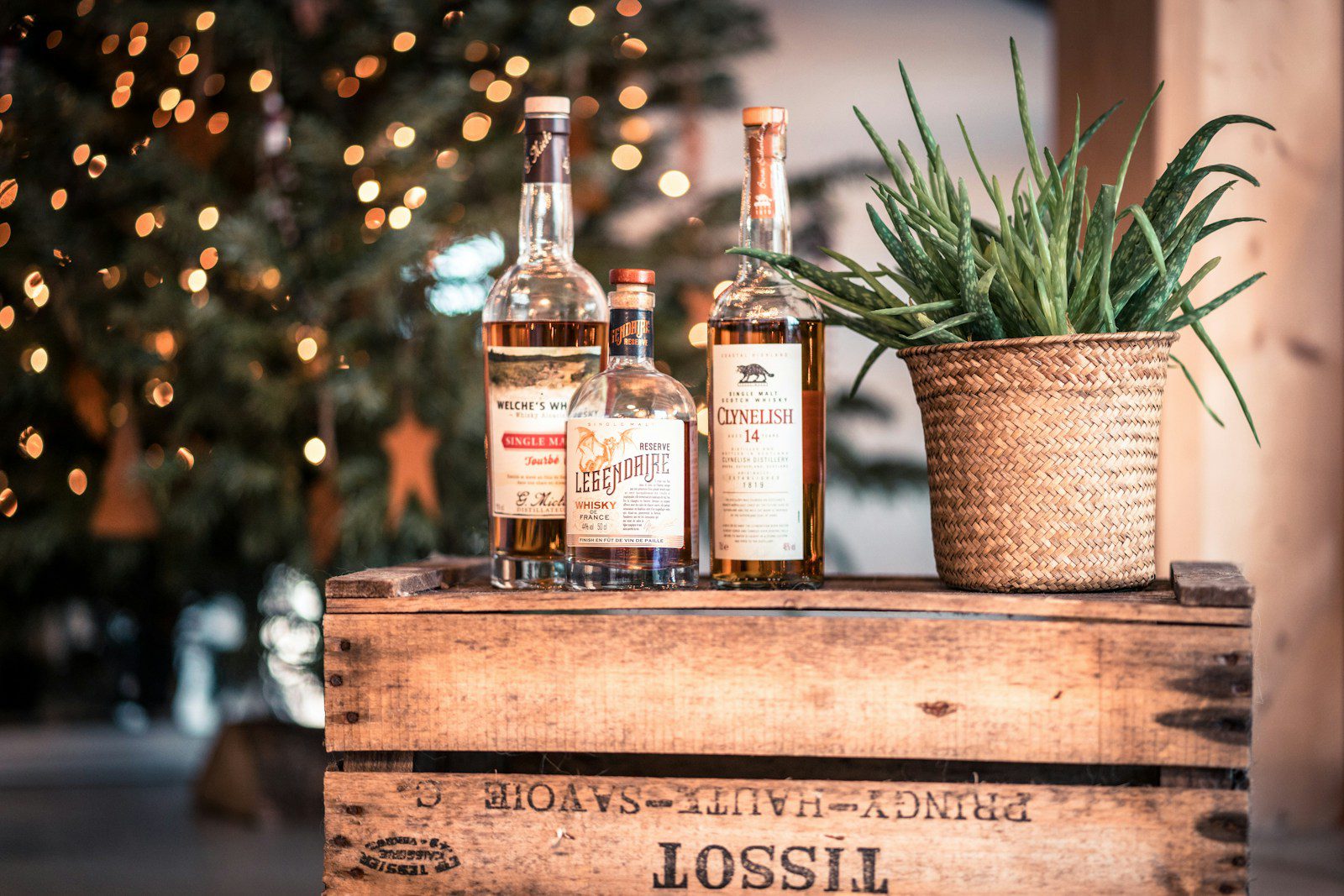 Trio of premium craft whisky bottles displayed on a rustic wooden crate with warm Christmas tree lights in the background. Curated holiday gift ideas for a boss.