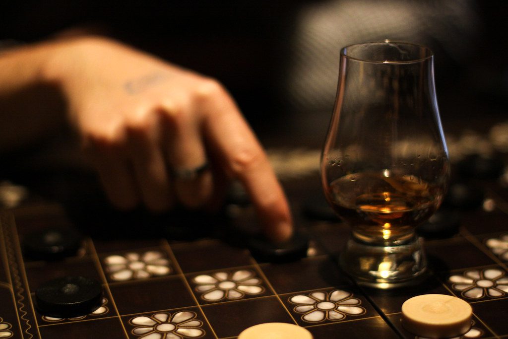 A close-up of a person's hand next to a Glencairn tasting glass filled with amber whisky, set against a dark background with a patterned board.