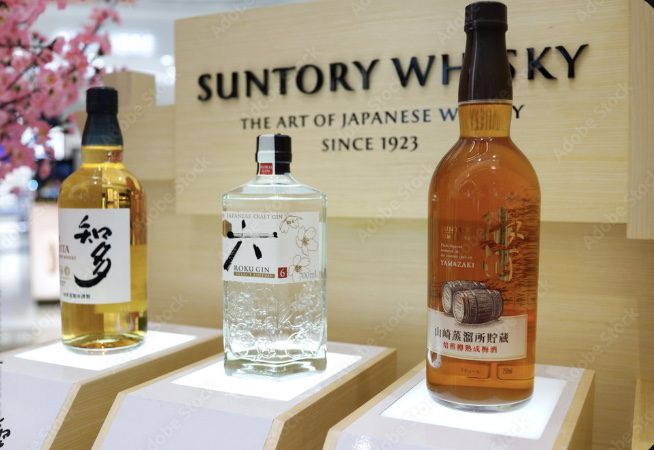 Three bottles of Japanese spirits displayed on a white stand under a "Suntory Whisky" sign. From left to right: a bottle of Chita Single Grain, a bottle of Roku Gin, and a limited-edition Suntory Old Whisky for 2026.