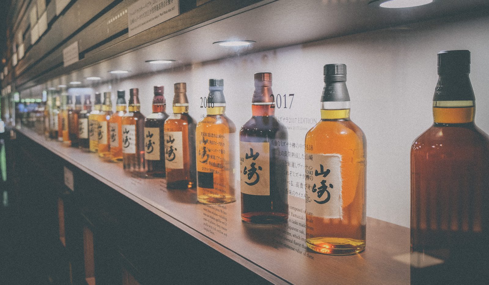 A row of Japanese whisky bottles on a wooden shelf, including Suntory and Nikka expressions, illuminated by soft overhead lighting.