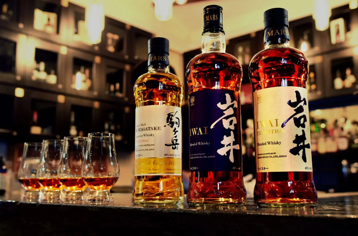 Three bottles of Mars Whisky standing on a dark bar counter next to five Glencairn tasting glasses filled with amber liquid. The bottles include Mars Komagatake Single Malt, Mars Iwai Blended Whisky (blue label), and Mars Iwai Tradition (gold label).