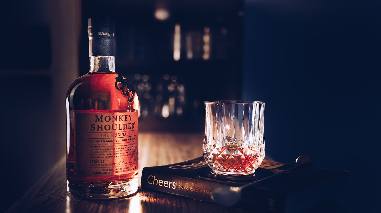 Monkey Shoulder whickey with a crystal glass filled with whiskey on a wooden bar counter. The range includes their signature scotch blended single malt whiskey expressions.